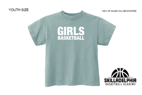 A youth-sized short-sleeve t-shirt in a dusty teal color with 'GIRLS BASKETBALL' printed in bold white letters on the front. The image includes 'YOUTH SIZE' text on the top left, '100% OF SALES WILL BE DONATED' on the top right, and the 'SKILLADELPHIA BASKETBALL ACADEMY' logo on the bottom right.