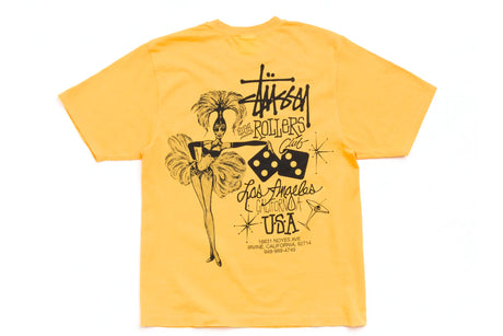 A yellow Stussy t-shirt with a black graphic print featuring a carnival dancer, dice, and 'Los Angeles Made in USA' text, displayed flat against a white background.