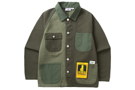 A color-block utility overshirt from Service Works in olive green and dark brown, featuring a button-up front, multiple utility pockets, and a yellow brand patch on the pocket.