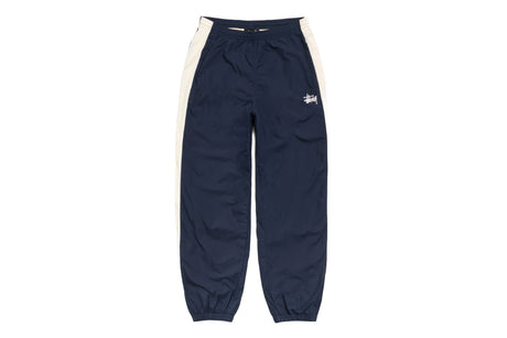 A pair of navy blue track pants with cream-colored side panels and a small white embroidered Stüssy logo on the left thigh, shown against a plain white background.