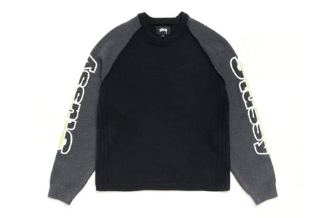 A Stussy raglan crewneck sweatshirt in black and dark gray, featuring the Stussy logo printed vertically in white on the sleeves with a neon green accent.